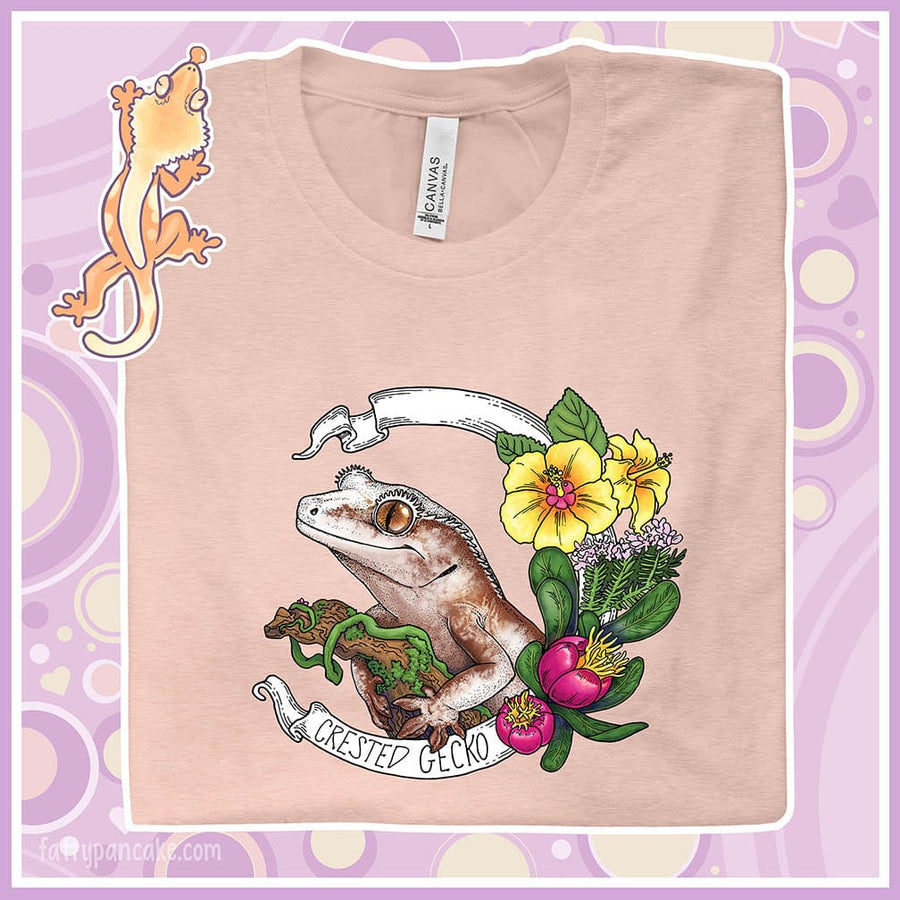 A folded peach colored crew neck tee showing an enlarged graphic of a crested gecko holding a branch with yellow hibiscus and other pink flowers as accents along the right of the design. A circular banner reads the name of the animal species on it.