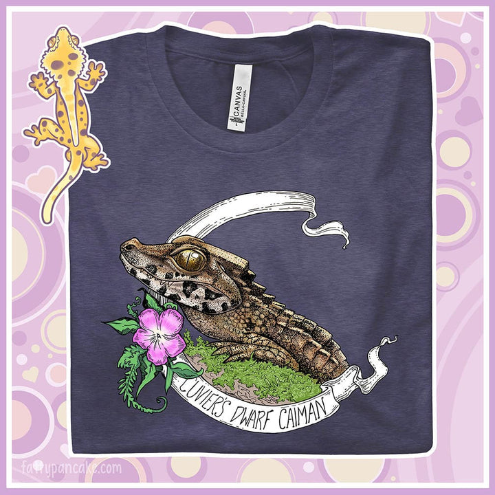 A folded heather midnight blue tee showing a Cuvier's Dwarf Caiman reptile perched on a mossy bed of greenery with a pink flower beneath it's head.