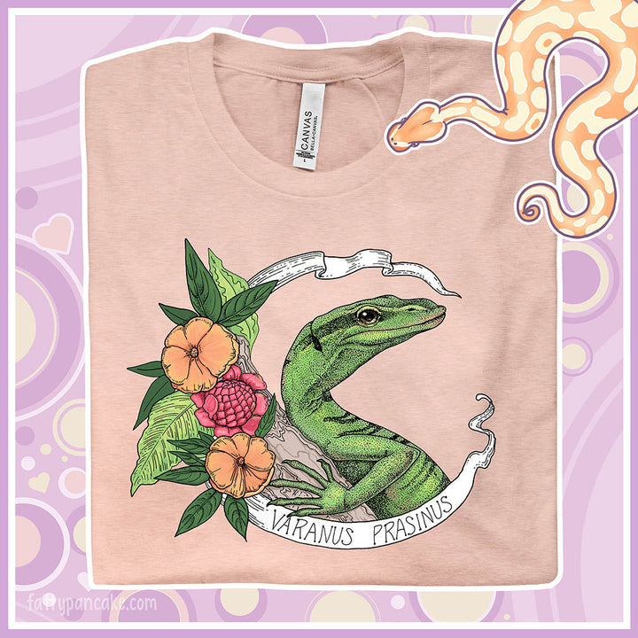 Folded peach color crew neck tee with a emerald tree monitor lizard in the center of a white banner with the species name on it and orange and pink flowers on the side.