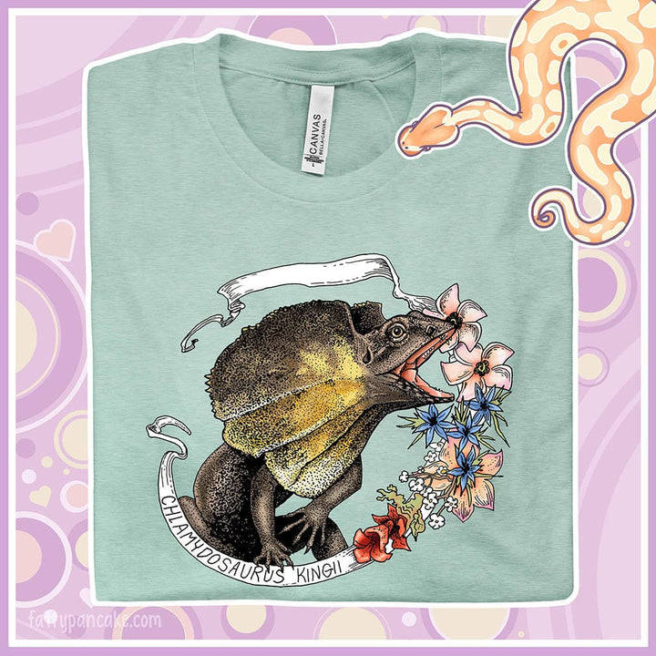 A folded dusty blue crew neck tee with a frilled lizard from Australia on it. It has a banner with it's species name and pink, blue, and rose colored flowers on the right side.