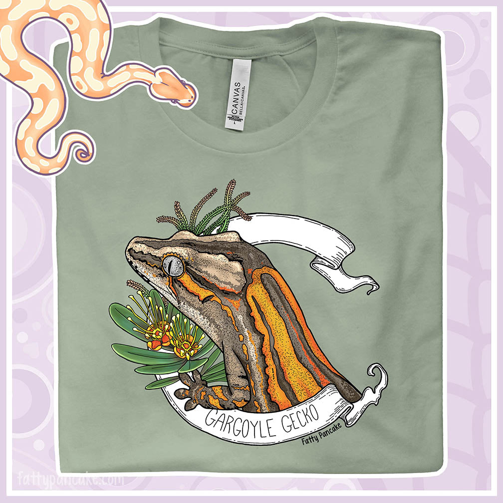 Green t-shirt with a gargoyle gecko design on a light purple background. A white banner frames the overall design with yellow and orange flowers and green leaves under the lizards head. The gargoyle gecko is tan with bright orange stripes.
