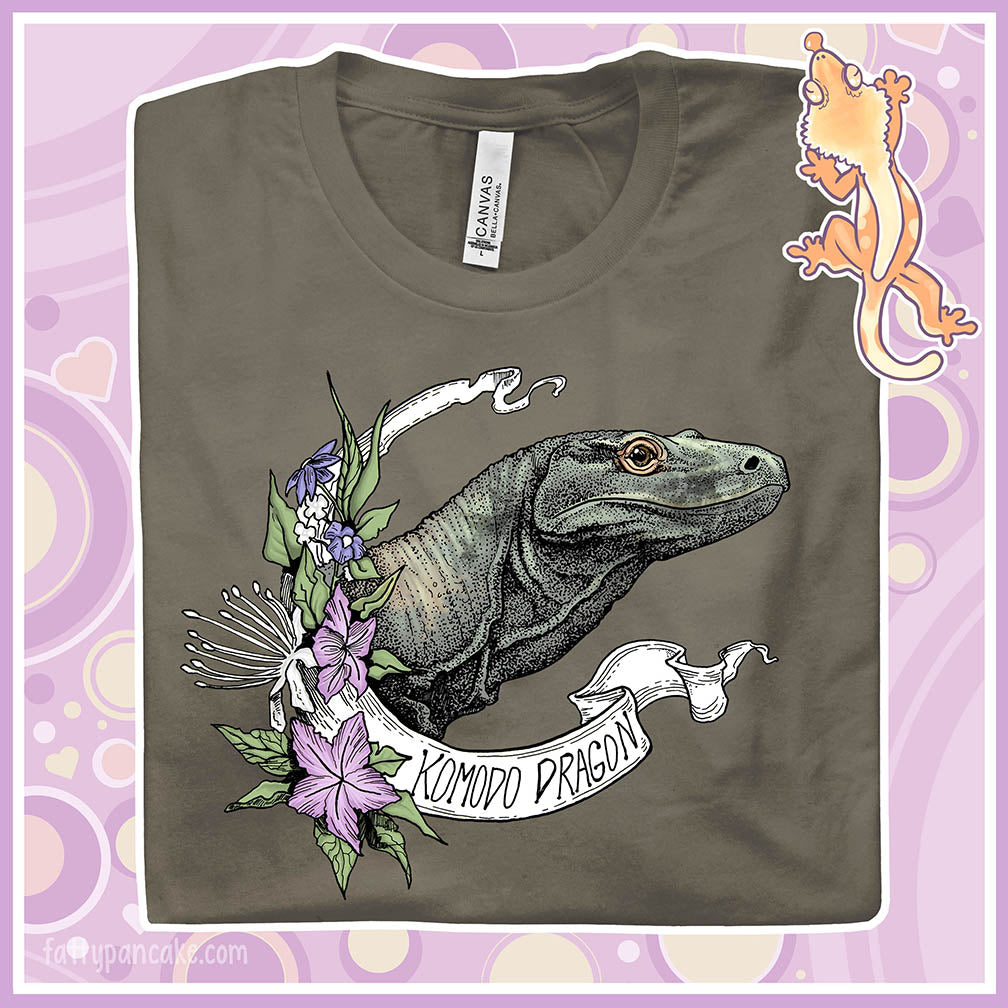 Folded tee showing an army colored crew neck tee with a head shot of a komodo dragon lizard framed by purple flowers and a white banner with text of the animal name.