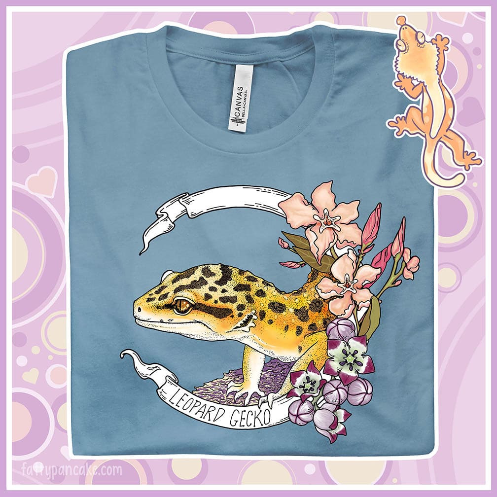 A folded steel blue crew neck tee with a leopard gecko art design printed on the front. A white banner encircles the reptile with the species name on it and pink and white flowers are on the right of the design.