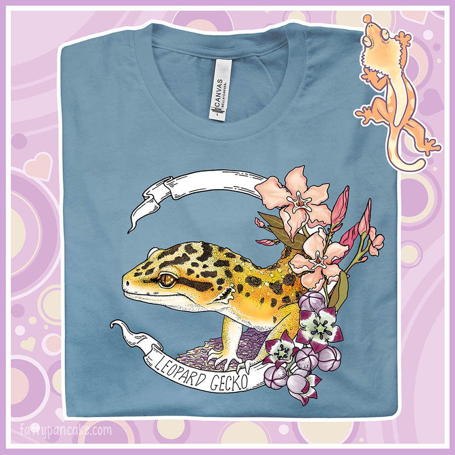 A folded steel blue crew neck tee with a leopard gecko art design printed on the front. A white banner encircles the reptile with the species name on it and pink and white flowers are on the right of the design.