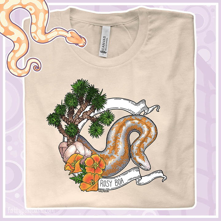 Beige t-shirt with a rosy boa illustration on a light purple background. The graphic has a white banner wrapping around the snake in a circle and has orange poppy flowers and a joshua tree on the left side of the graphic. The name Rosy Boa is on the bottom part of the banner.
