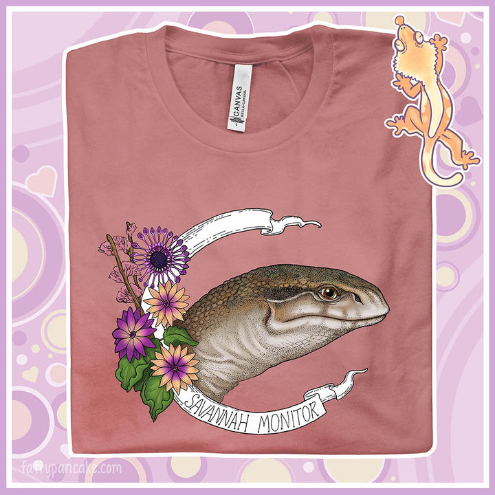 A folded mauve crew neck tee with a head shot of a savannah monitor lizard with purple flowers on the side and a white banner encircling the design which says the species name.