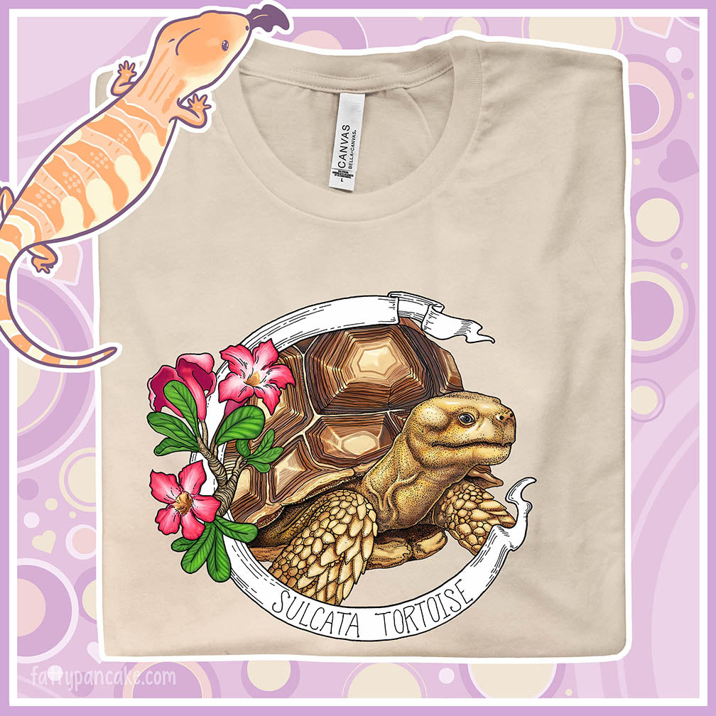 A folded soft cream colored crew neck tee shirt with a sulcata tortoise in the middle of a white banner and pink flowers on the left side of the design. The banner reads Sulcata Tortoise.