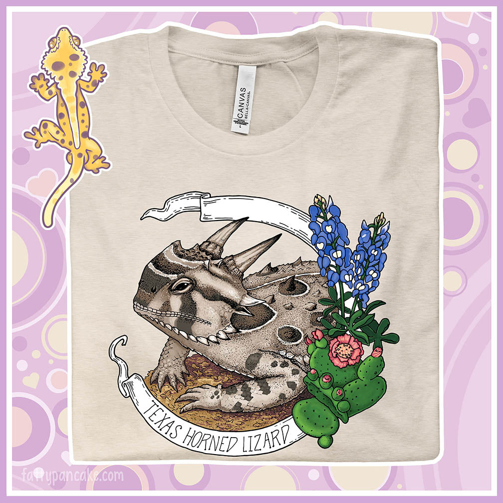 A folded heather dusty colored crew neck tee with a texas horn toad lizard resting on sand and framed by cactii and blue bonnet flowers. A banner encircles the design with the text "texas Horned Lizard" on it.