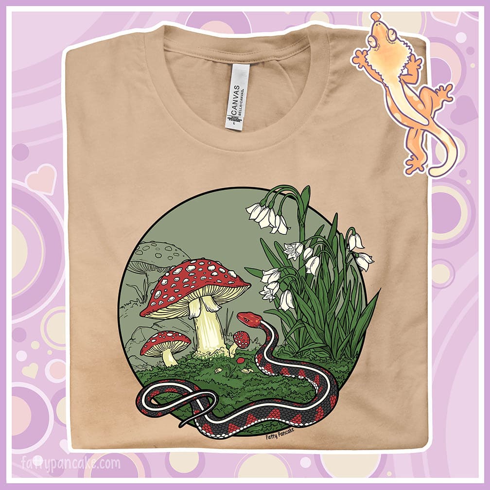 Image depicts a folded crew neck tee so the graphic is enlarged. The design is a California red sided garter snake coiled up near some mushrooms and white bell flowers. It has a very neutral color palette with red capped mushrooms and simple 5 color design.