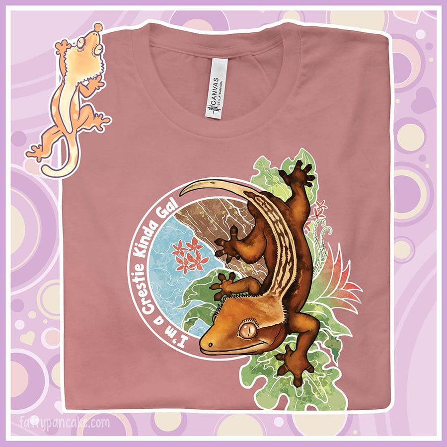 A folded crew neck tee showing an enlarged image of a graphic of a crested gecko climbing down from a tree with the words "I'm a crestie kind of gal" on the left hand side.