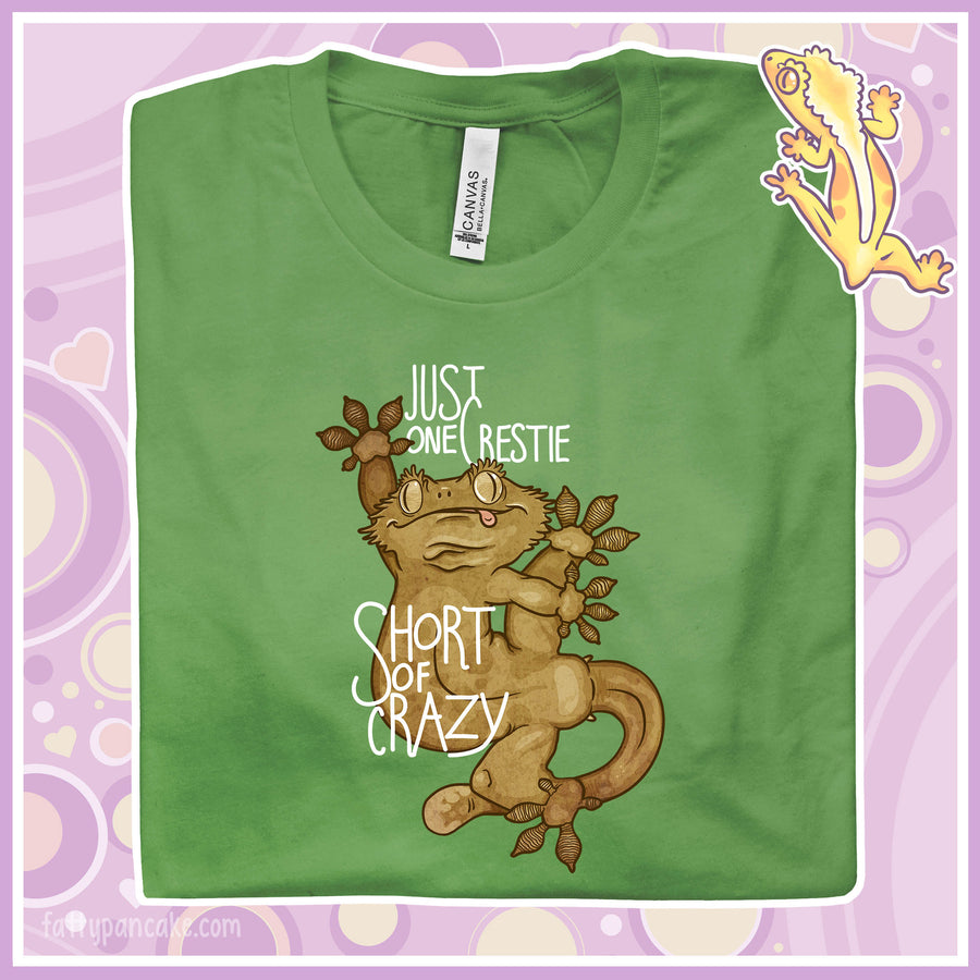 A folded green crew neck tee with the words "Just one Crestie Short of Crazy" with a crested gecko design clinging to the center. It's legs are flung out and it has a silly expression.