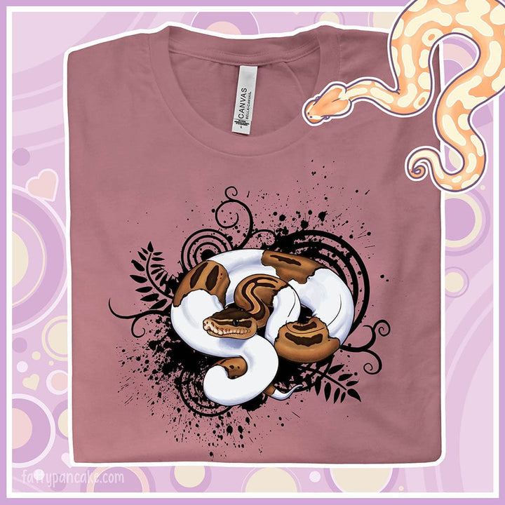 Mockup image showing a crew neck tee folded face up with a pied ball python coiled up on solid black spatters, swirl designs, and leaves. The background is purple with hearts and circles and a cute cartoon ball python is on the corner.