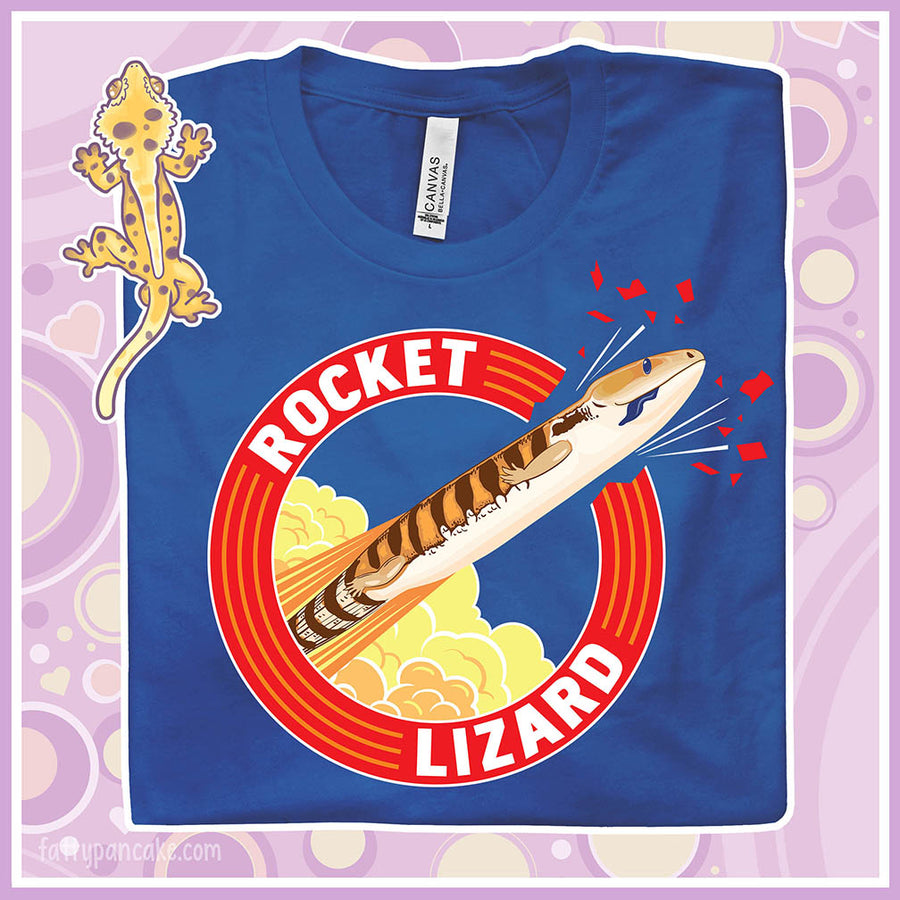 A folded blue crew neck tee with a circular design stating Rocket Lizard on the top and bottom and a cartoon blue tongue skink lizard blasting out from the side.