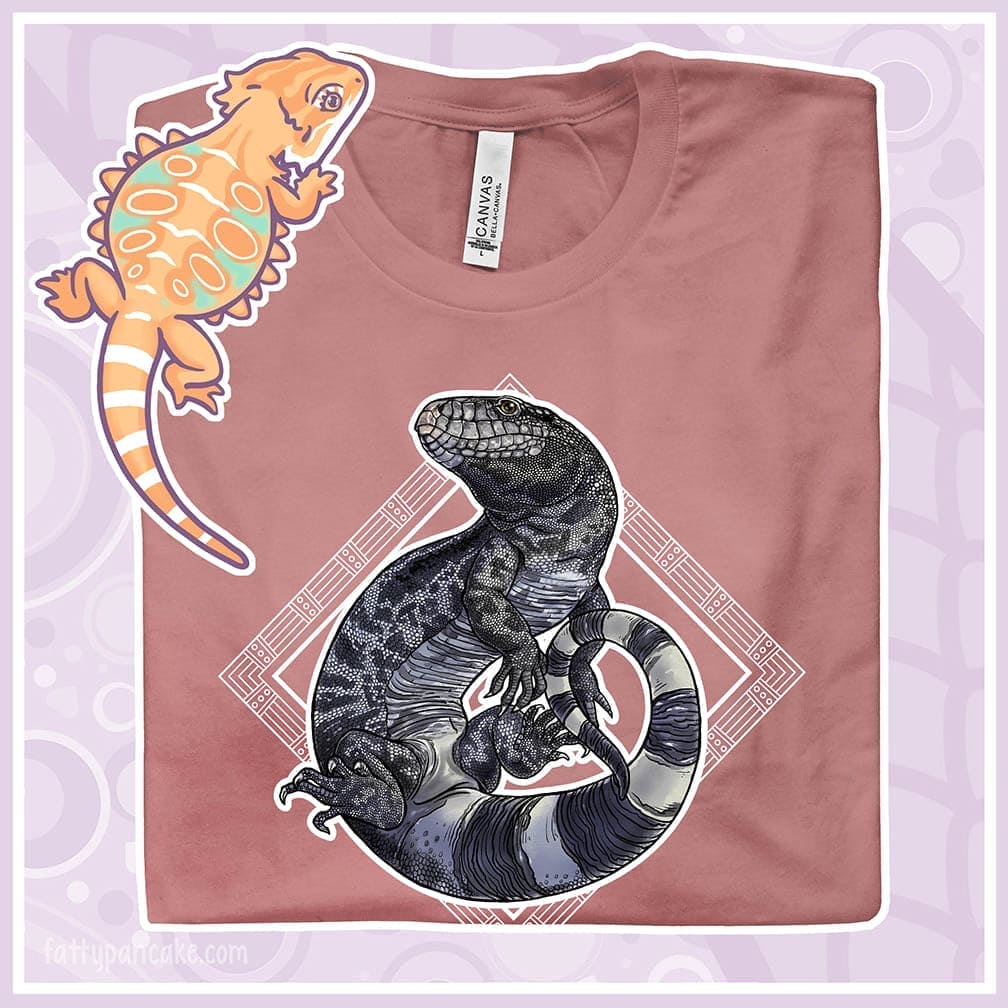 Pink t-shirt with a black and white tegu lizard graphic and a colorful lizard illustration on a light purple background.