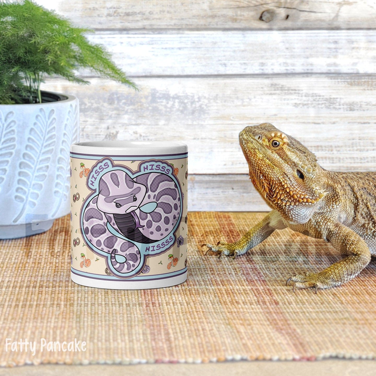 A white ceramic mug with a purple and gray snake design. The mug has the word 'HISS' written on it three times. The mug is sitting on a brown woven mat. A bearded dragon lizard is looking at the mug.