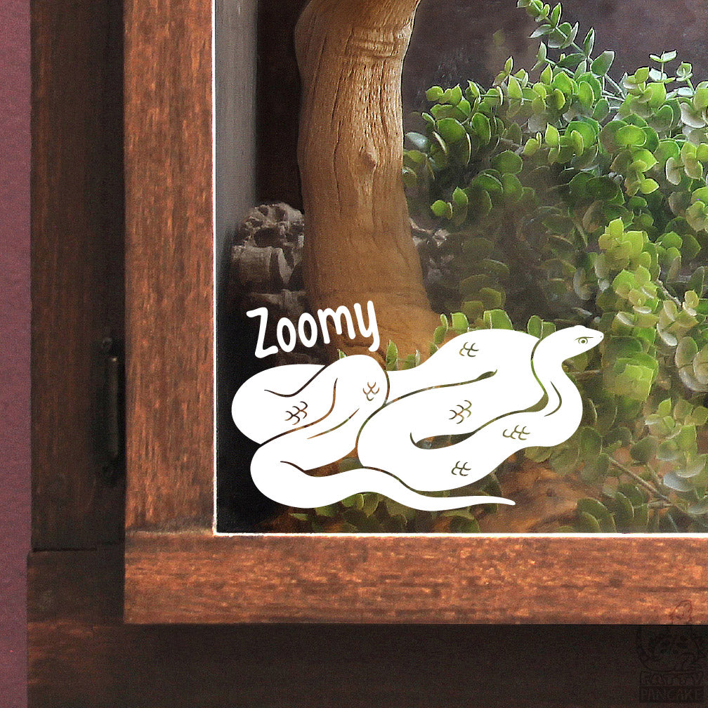 A close up of a wooden reptile enclosure with a white vinyl decal of a Black Racer snake with the name Zoomy above it's shell. The interior of the enclosure has greenery and branches.
