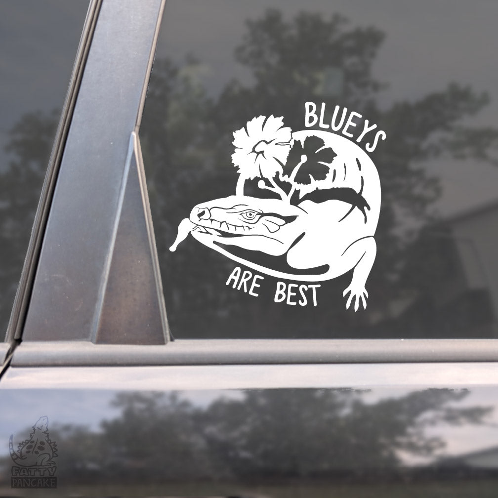A White vinyl decal of a blue tongue skink with 2 flowers and the text "Blueys are Best" framing the design placed on the window of a car.