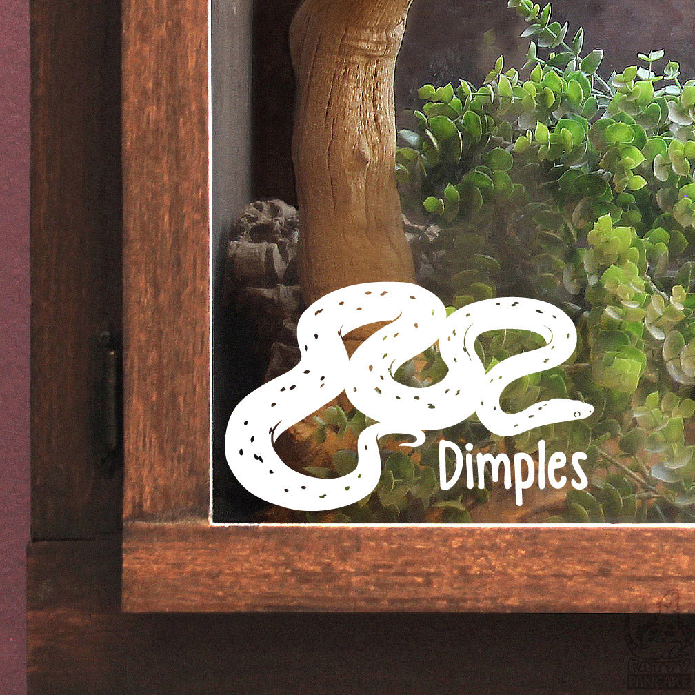 A white vinyl decal of a coiled Palmetto morph Corn Snake snake with the name 'Dimples' printed next to it, shown applied to the glass door of a wooden terrarium.