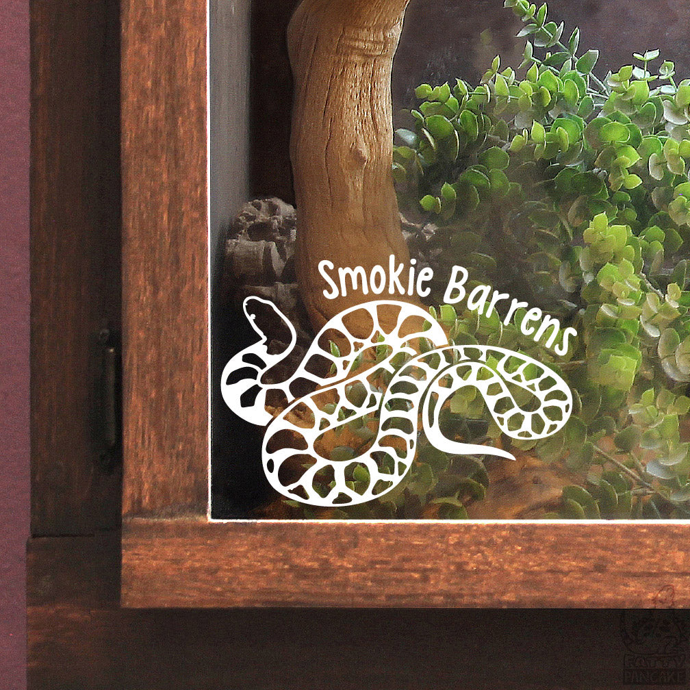 A white vinyl decal of a coiled Desert Kingsnake with the text 'Smokie Barrens' printed on it, shown applied to the glass door of a wooden display case containing a live snake and green plants.