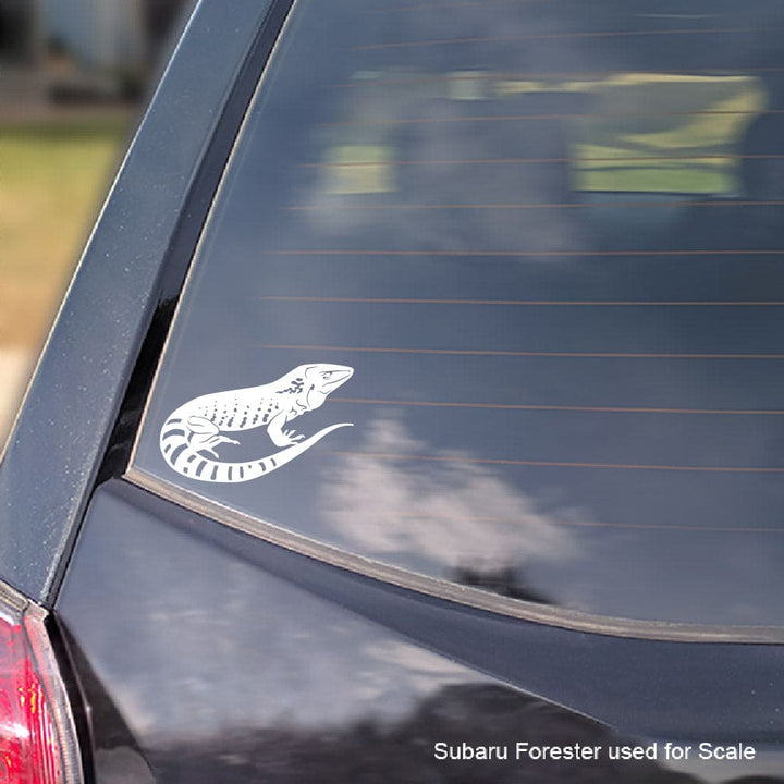 A white vinyl decal of a Dusky Giant Ameiva lizard applied to the rear window of a dark-colored car, shown for scale with a Subaru Forester watermark in the bottom right corner.