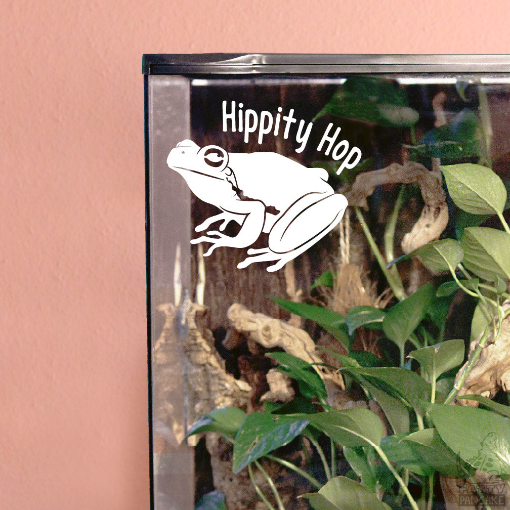 A white vinyl decal featuring a frog and the text 'Hippity Hop' applied to the glass of a terrarium, shown against a pink wall background.