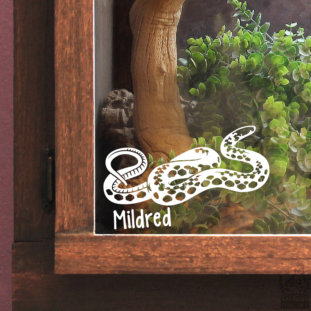 A white vinyl decal of a coiled Kirtland's Snake with the name 'Mildred' printed below it, applied to the glass front of a wooden terrarium containing green plants and a wood branch.