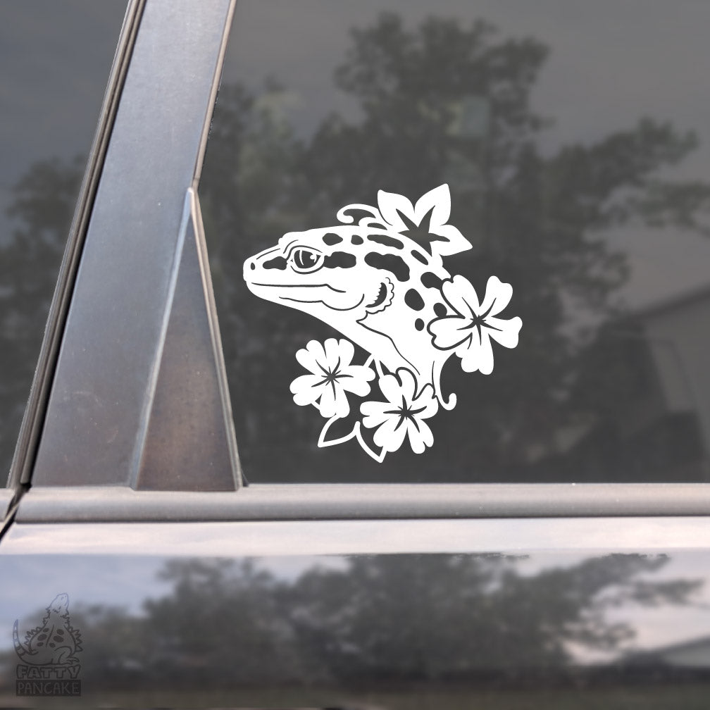 Image shows a close up of a car window with an elegant looking white vinyl decal of a Leopard Gecko lizard facing left with four flowers framing it. 