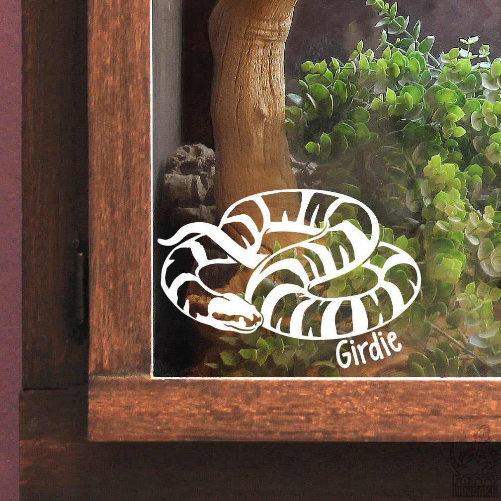 A white vinyl decal of a coiled Puffed-Faced Water Snake with the name 'Girdie' written below it, shown applied to the glass front of a wooden reptile terrarium containing a wooden branch and green plants.