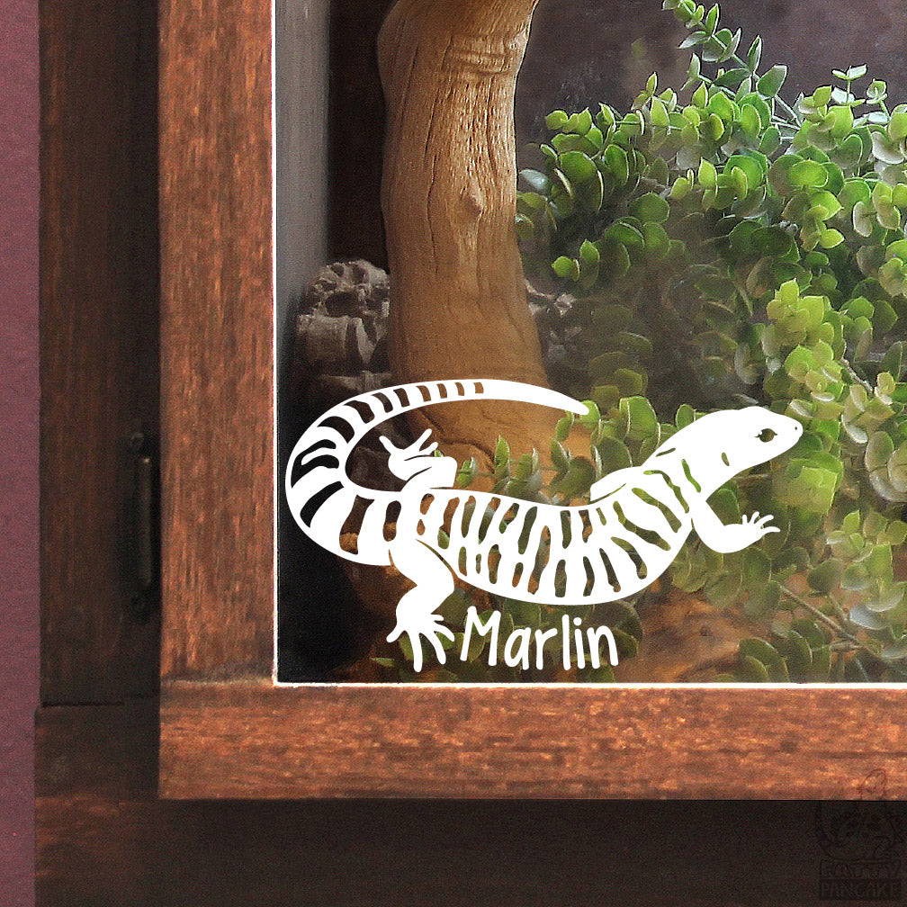 A white vinyl decal of a Schneider's Skink lizard with the name 'Marlin' printed underneath, shown applied to the glass door of a wooden terrarium containing a wood leg and green plants.
