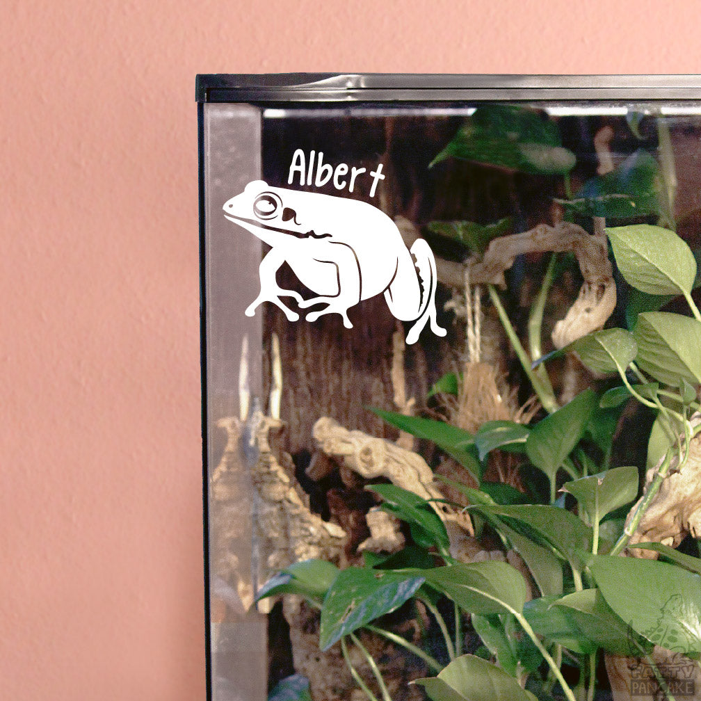 A white vinyl decal of a White Lipped frog with the name 'Albert' written above it, applied to the glass front of a terrarium containing green plants and wood branches, set against a pink wall.