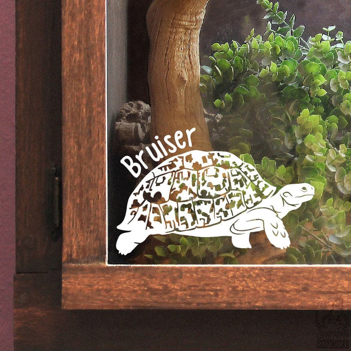 A white Leopard Tortoise decal with the name 'Bruiser' on a glass panel with greenery inside the wooden enclosure.