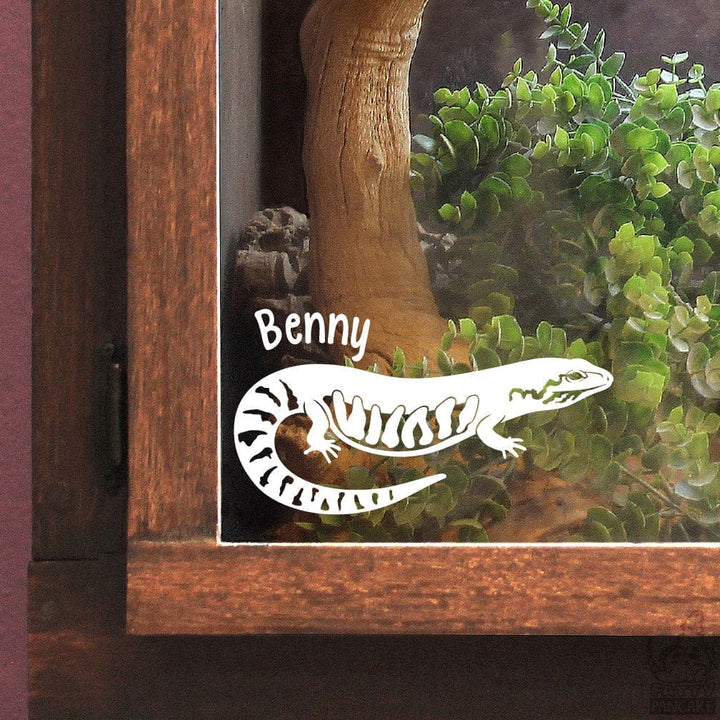 Decorative fire skink lizard decal named 'Benny' on a glass panel with a wooden frame enclosure and plant background.