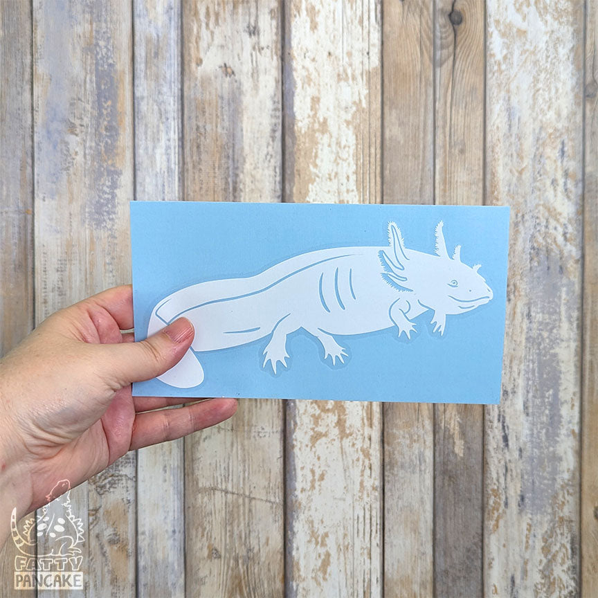 A hand holds a rectangular light blue paper featuring a white axolotl  decal, set against a rustic whitewashed wood plank background.