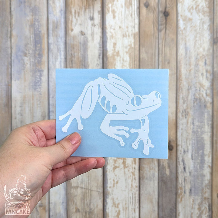 A hand holds a rectangular light blue paper featuring a white Red eyed Tree Frog decal, set against a rustic whitewashed wood plank background.
