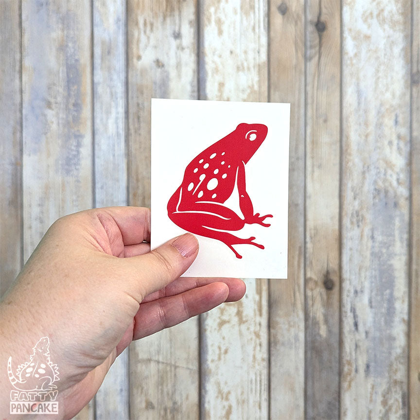 A hand holds a rectangular white paper featuring a red Spotted Poison Dart Frog decal, set against a rustic whitewashed wood plank background.