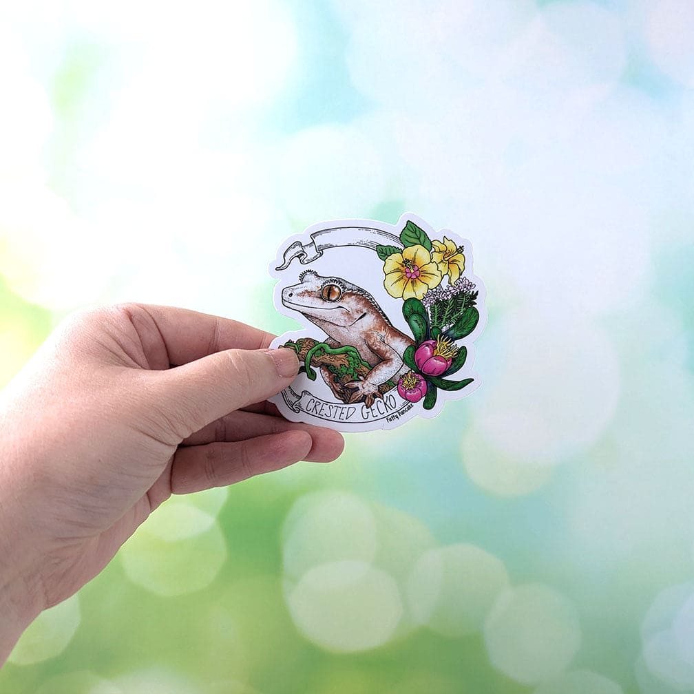 A hand holding a sticker featuring a crested gecko with a brown and white body, perched on a branch with green moss. The gecko is surrounded by yellow hibiscus flowers, pink flowers, and green leaves.