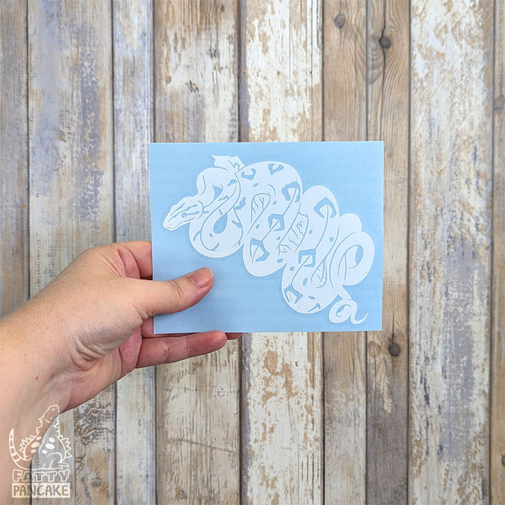 A hand holds a rectangular light blue paper featuring a white Amazon Tree Boa decal, set against a rustic whitewashed wood plank background.