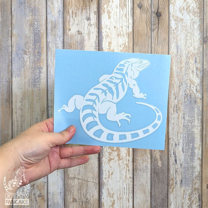 A hand holds a light blue square card featuring a white decal design of a tegu lizard, set against a rustic wooden plank background.