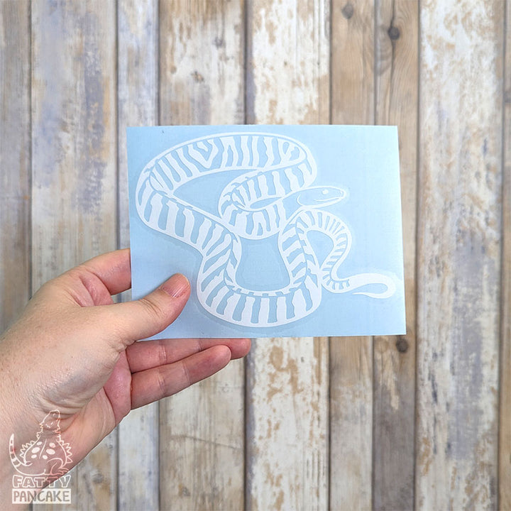 A hand holds a rectangular light blue paper featuring a white Black Headed Python decal, set against a rustic whitewashed wood plank background.