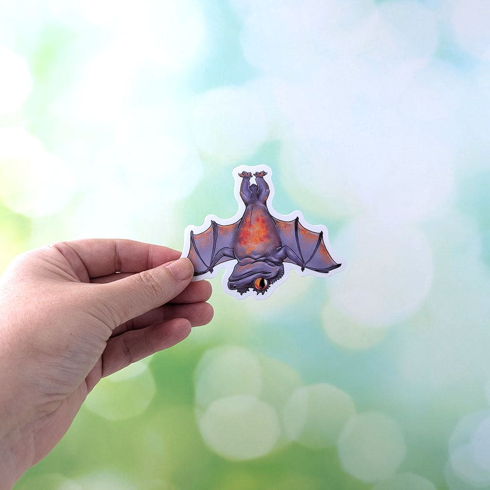 A hand holding a Halloween themed sticker of a crested gecko bat character for scale.