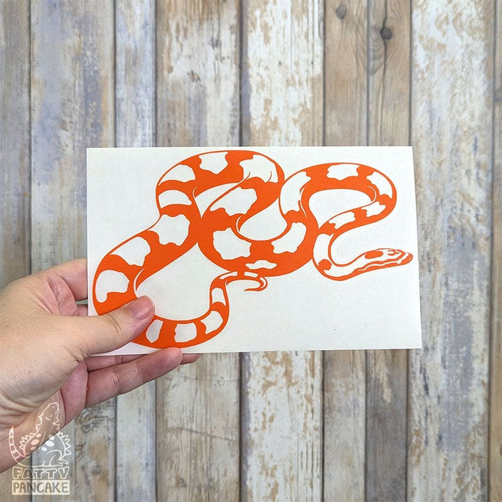 A hand holds a white rectangular sticker or card featuring a bold orange corn snake graphic, set against a rustic light wood plank background.