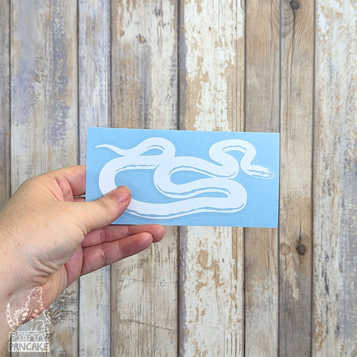 A hand holds a rectangular light blue paper featuring a white African House Snake decal, set against a rustic whitewashed wood plank background.