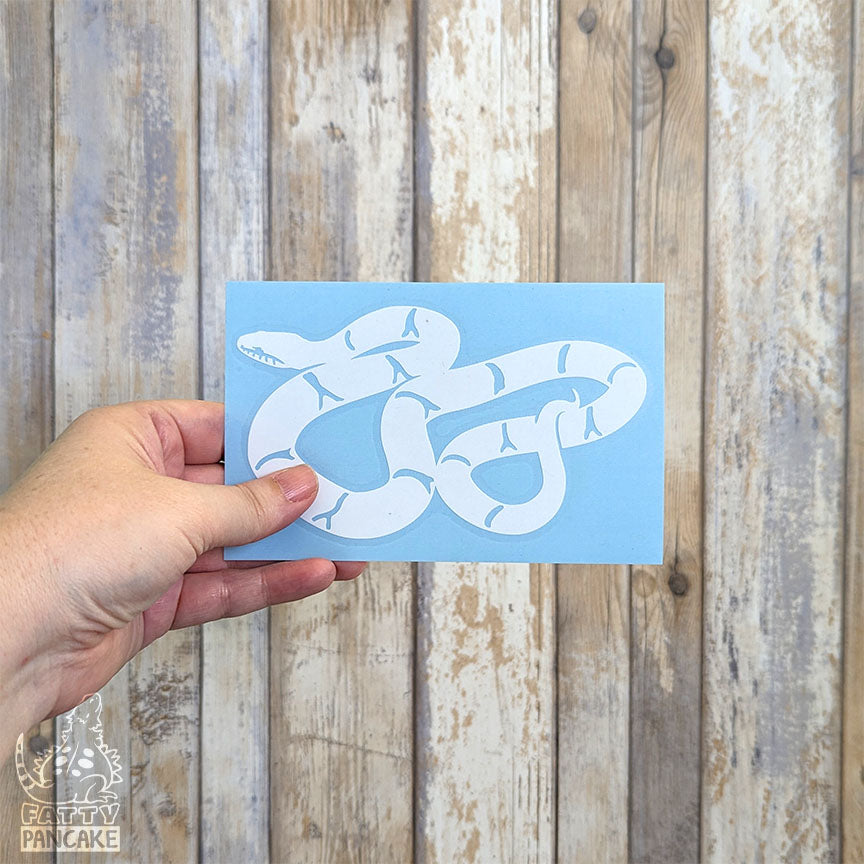 A hand holds a rectangular light blue paper featuring a white Russian Rat Snake decal, set against a rustic whitewashed wood plank background.