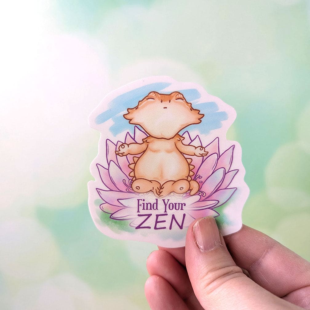 Hand holding vinyl sticker of a cartoon bearded dragon meditating on a lotus flower and the words, "Find your Zen" underneath them. 
