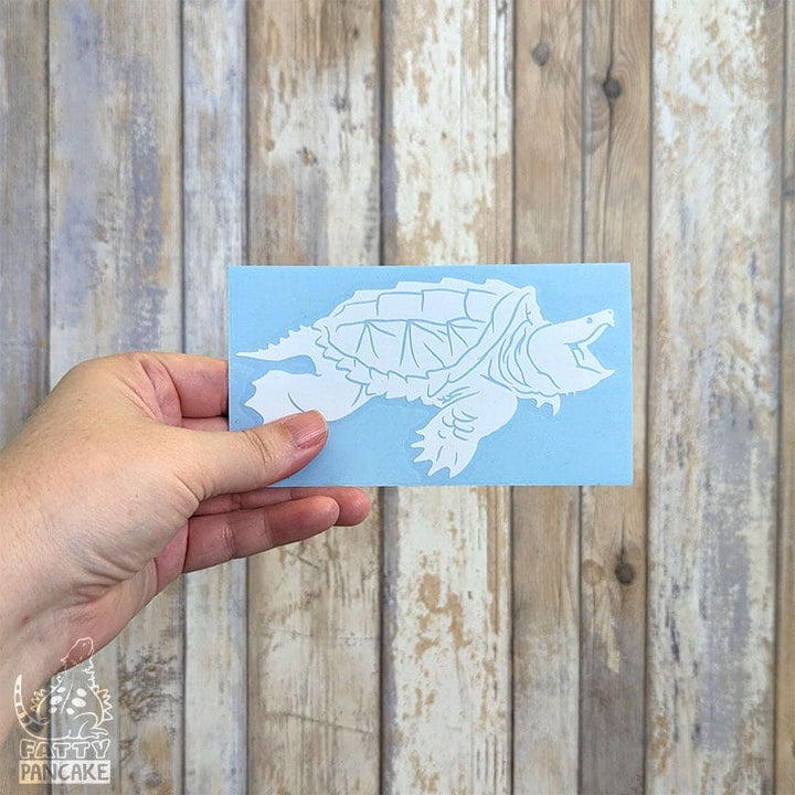A hand holds a rectangular light blue card featuring a white alligator snapping turtle decal, set against a rustic whitewashed wood plank background. 