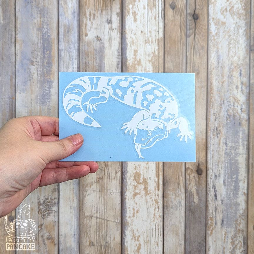 A hand holds a rectangular light blue paper featuring a white Gila Monster decal, set against a rustic whitewashed wood plank background.