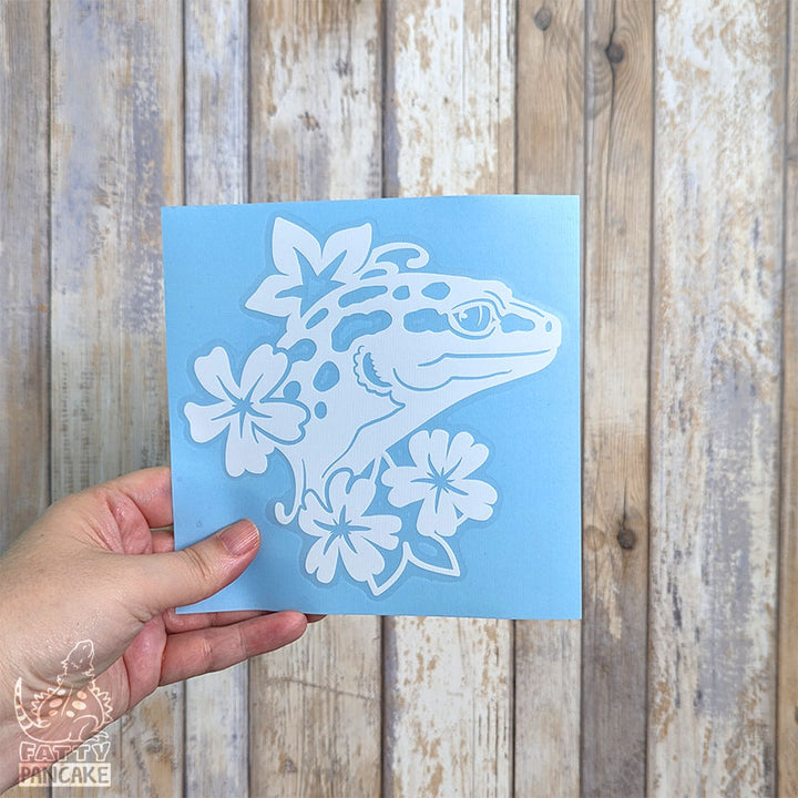 A hand holds a square light blue paper featuring a white die-cut design of a leopard gecko surrounded by flowers, set against a rustic wooden plank background.