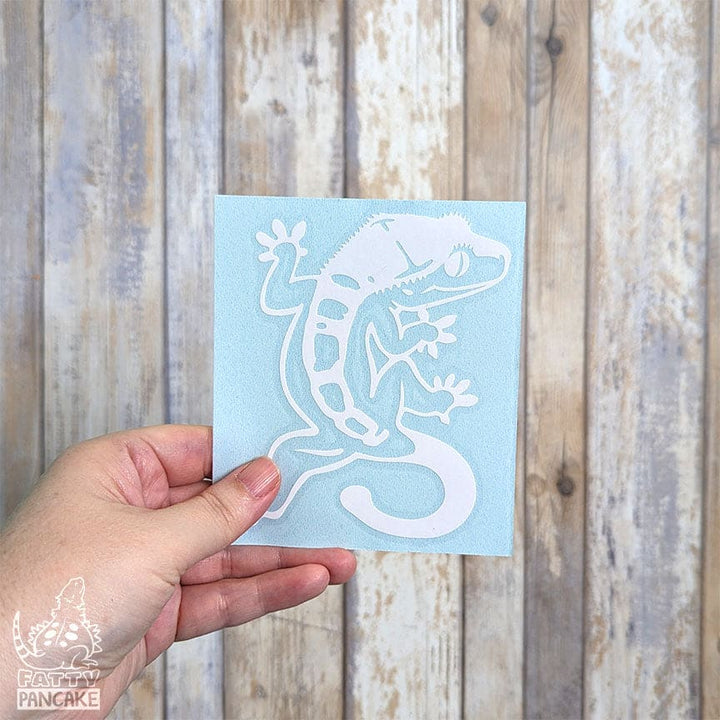 A hand holds a small square light blue card featuring a white gecko decal, set against a rustic whitewashed wood plank background. 