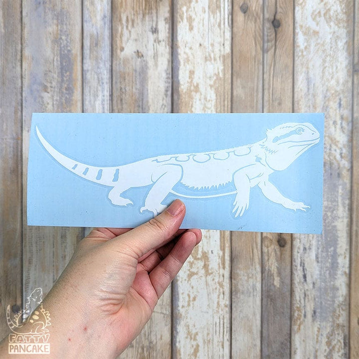 A hand holds a light blue rectangular sticker or decal featuring a white bearded dragon lizard decal, set against a rustic weathered wood plank background.