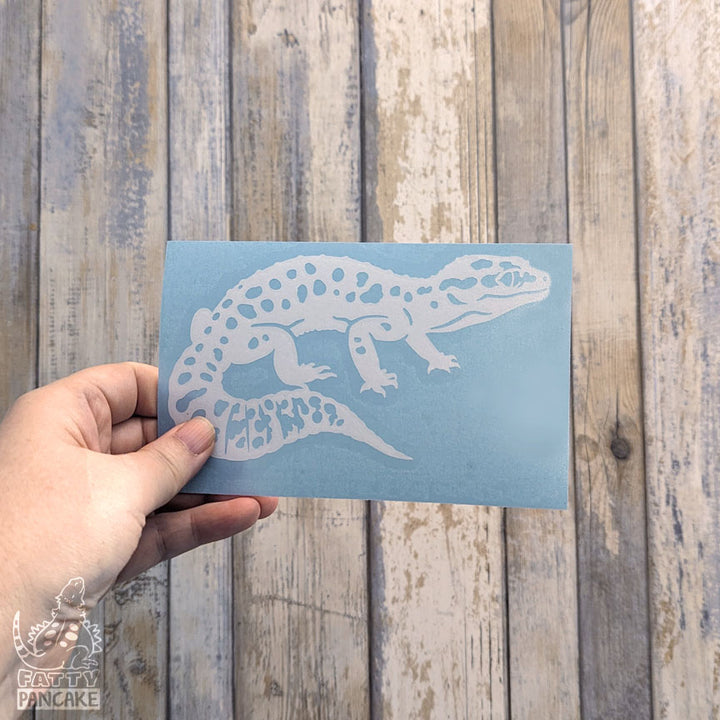 A hand holds a rectangular light blue paper featuring a white Leopard Gecko decal, set against a rustic whitewashed wood plank background.